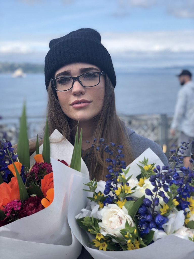 Morgan in glasses and a beanie holding colorful bouquets beside the water